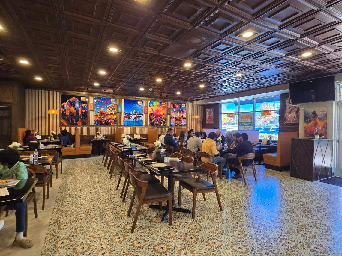 The spacious dining room has a richly patterned floor, dark wood tables, and a coffered ceiling. Large, colorful posters hang on the walls, and several groups of people are seated and dining.