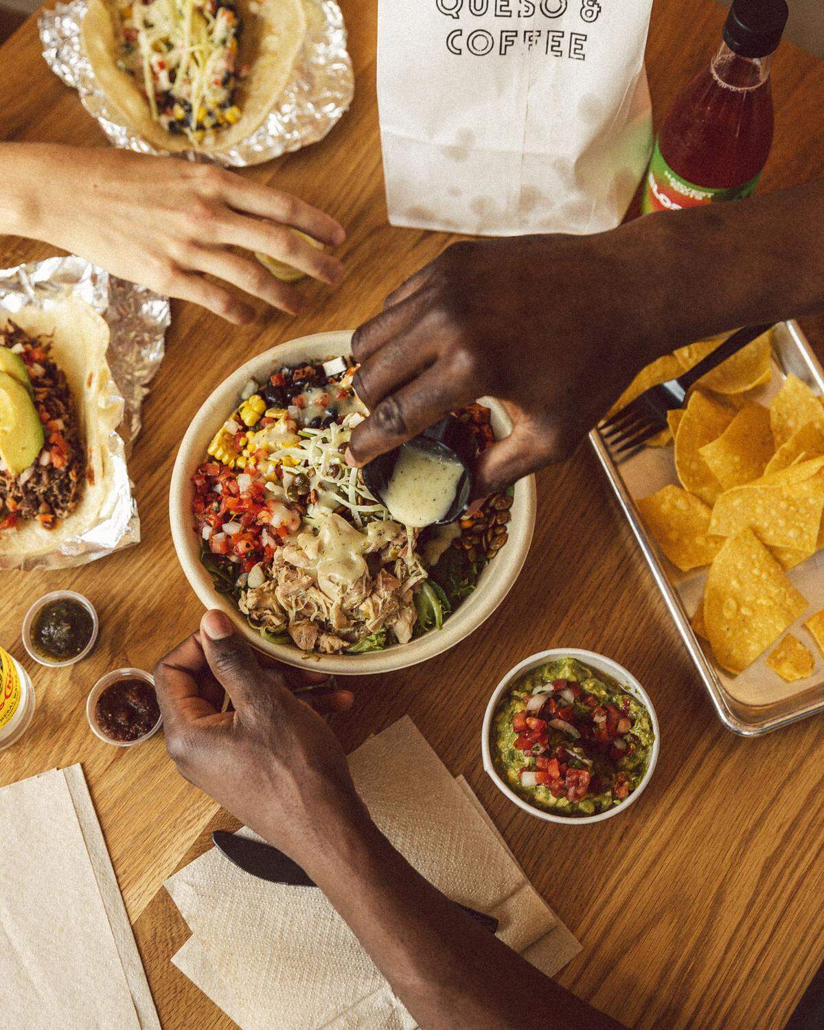 A top-down shot of a wooden table spread with a colorful burrito bowl, various tacos, chips, and guacamole. Two people are sharing the meal, with one person pouring a light green dressing over the central bowl filled with chicken, corn, and black beans.