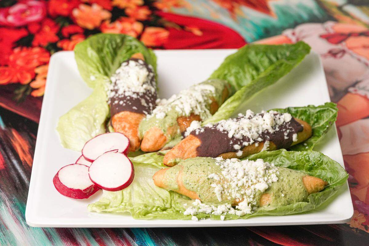 Four golden-fried molotes are nestled in individual Romaine lettuce leaves and topped with a variety of green and dark red salsas and crumbled white cheese. Three thin slices of bright red radish sit on the side of the white square plate as a fresh garnish. The dish is presented on a colorful floral tablecloth that adds a vibrant background to the meal.
