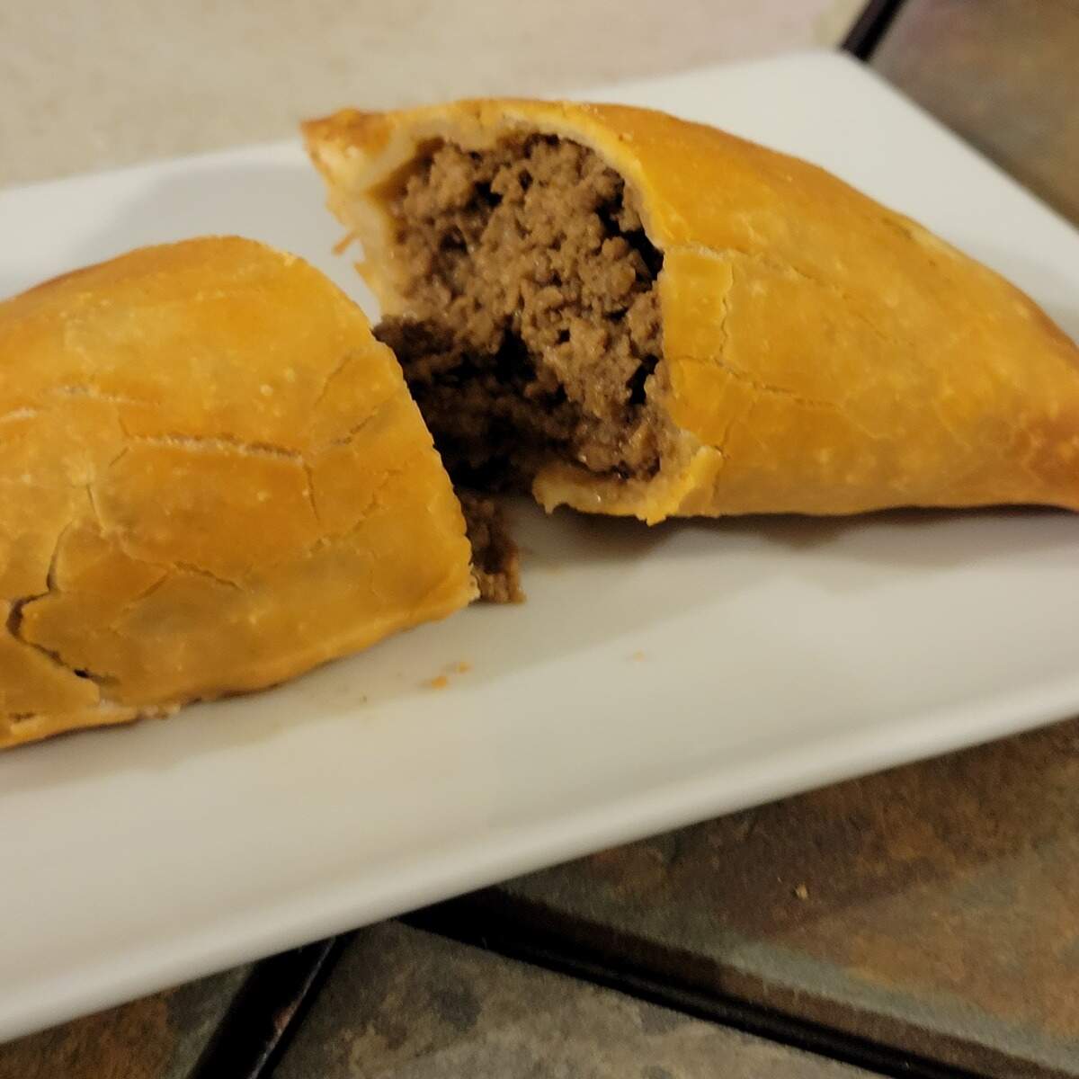 A baked empanada on a white plate is cut open, showing a savory ground-meat filling. The golden crust is cracked and slightly flaky.