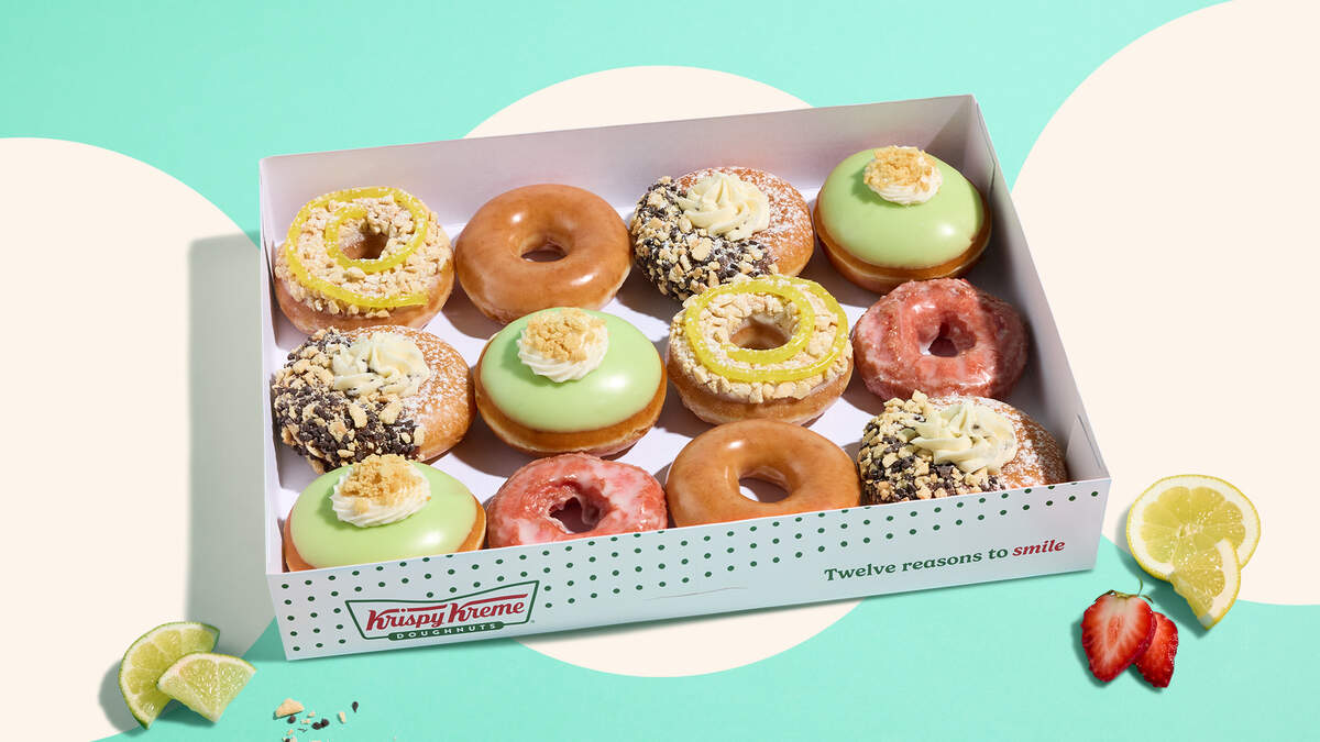 An open white Krispy Kreme box filled with a dozen assorted doughnuts sits against a teal and cream patterned backdrop. The collection showcases classic original glazed doughnuts alongside festive summer varieties featuring pink, light green, and lemon-yellow icings, crumbles, and whipped cream toppings. Fresh lime wedges, lemon slices, and a cut strawberry are artfully arranged on the surface outside the box, which bears the slogan “Twelve reasons to smile.”