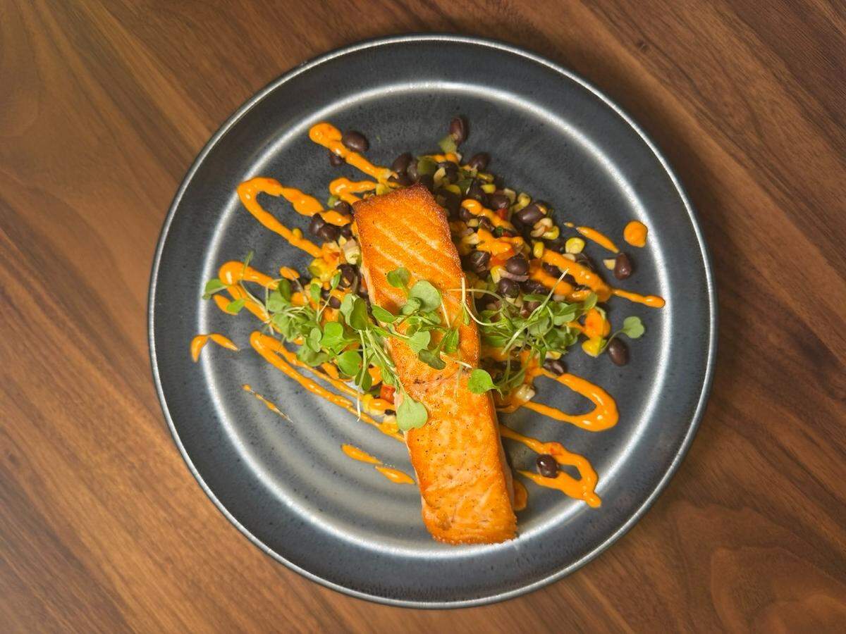 A perfectly seared salmon fillet is presented on a dark slate-gray plate, resting atop a colorful bed of black beans and corn salsa. Bright orange crema is artistically drizzled across the entire dish in diagonal lines, accented by a delicate garnish of fresh microgreens. The plate is photographed from an overhead perspective against a warm, natural wood tabletop that highlights the vibrant colors of the meal.