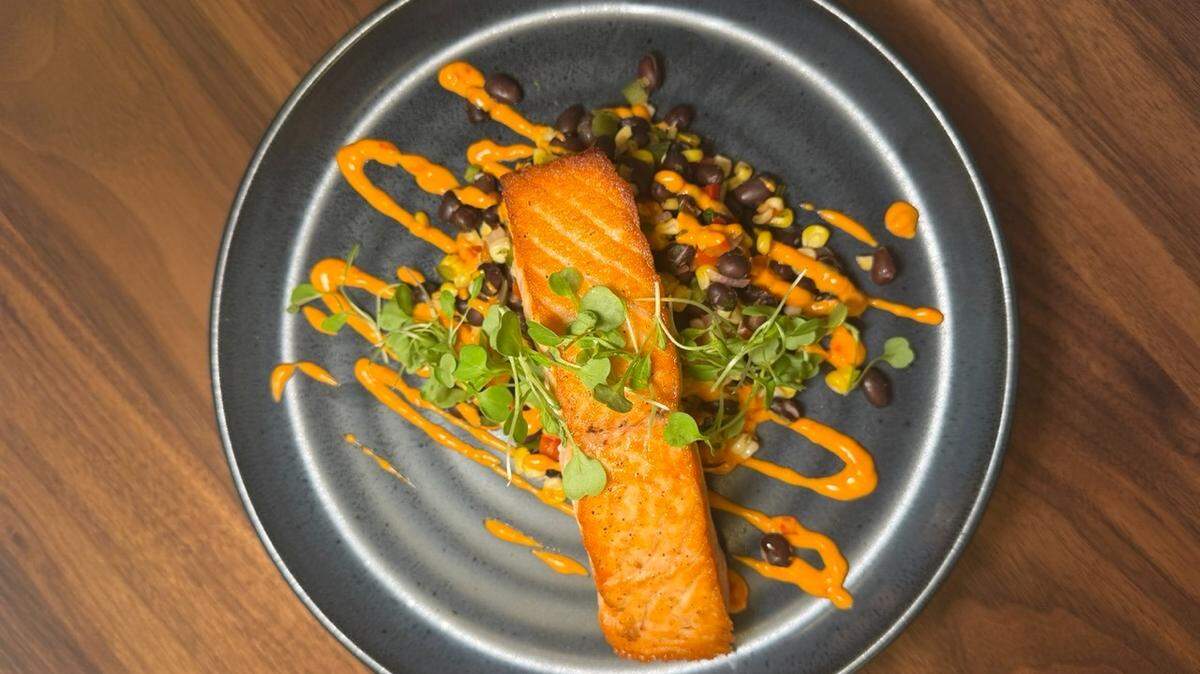 A perfectly seared salmon fillet is presented on a dark slate-gray plate, resting atop a colorful bed of black beans and corn salsa. Bright orange crema is artistically drizzled across the entire dish in diagonal lines, accented by a delicate garnish of fresh microgreens. The plate is photographed from an overhead perspective against a warm, natural wood tabletop that highlights the vibrant colors of the meal.