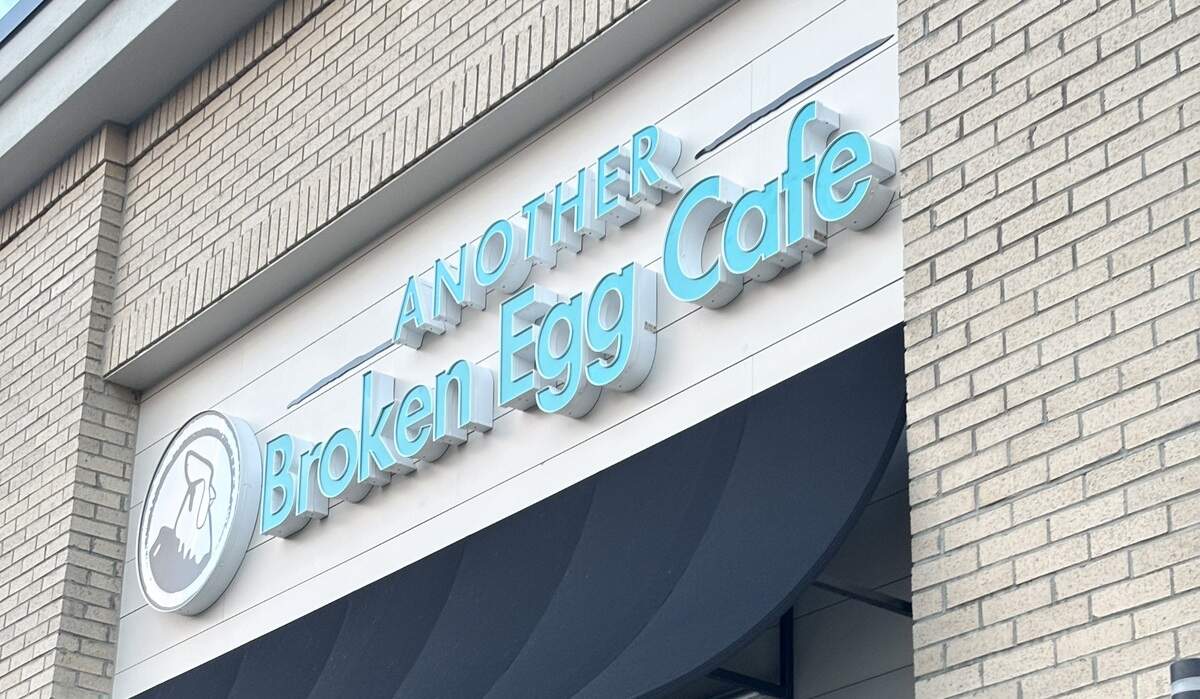 An outdoor, angled close-up shows the storefront sign for “Another Broken Egg Cafe” mounted on a white surface between light-colored brick walls. The restaurant’s name features bright turquoise channel letters, with a circular white logo displaying a stylized rooster to the left. A black scalloped awning hangs directly below the signage.