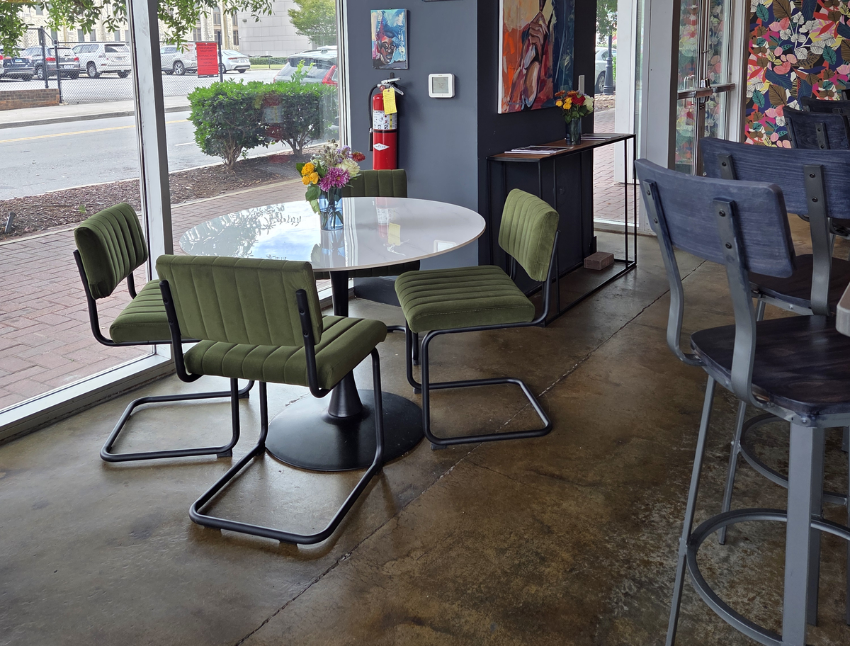 This photo shows a cafe interior with large floor-to-ceiling windows looking out onto a street. A small white table is surrounded by four modern, olive-green upholstered chairs, with a colorful flower arrangement on top of the table.