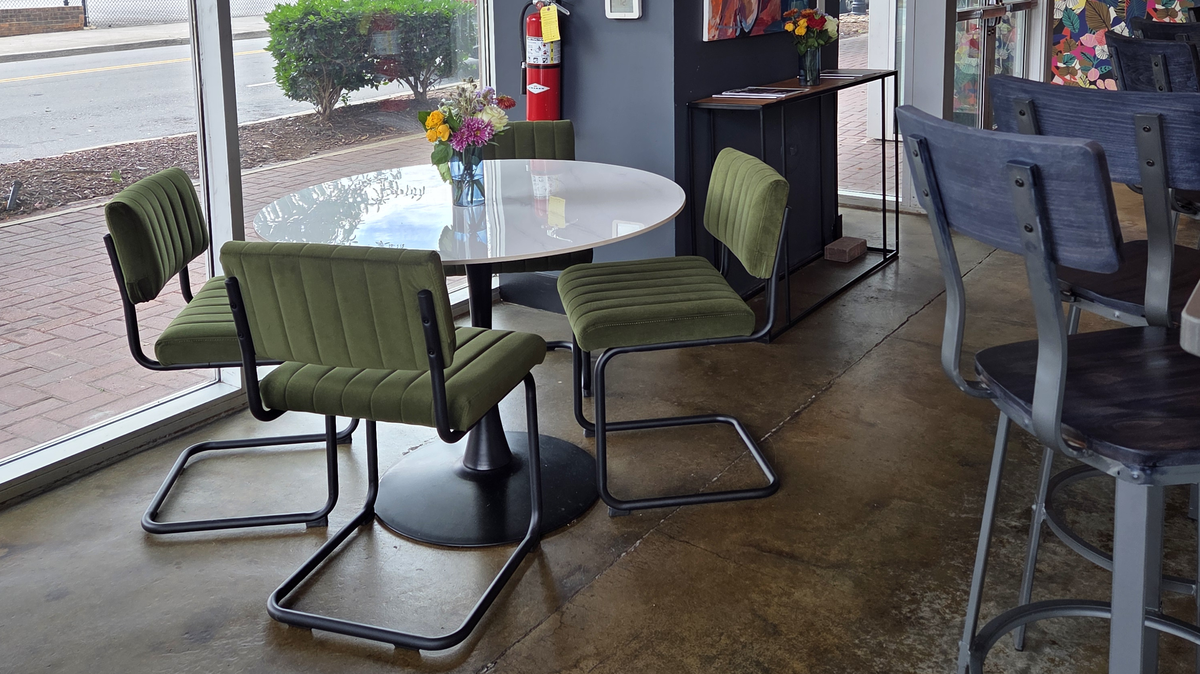 This photo shows a cafe interior with large floor-to-ceiling windows looking out onto a street. A small white table is surrounded by four modern, olive-green upholstered chairs, with a colorful flower arrangement on top of the table.