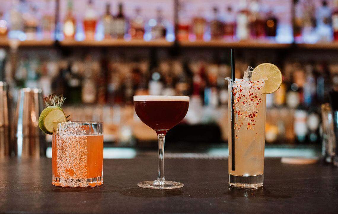 Three distinct, colorful cocktails are lined up on a dark bar top in front of a brightly lit display of liquor bottles. The drinks include one with a salt-rimmed glass, one in a coupe, and a tall one with chili flakes on the rim.