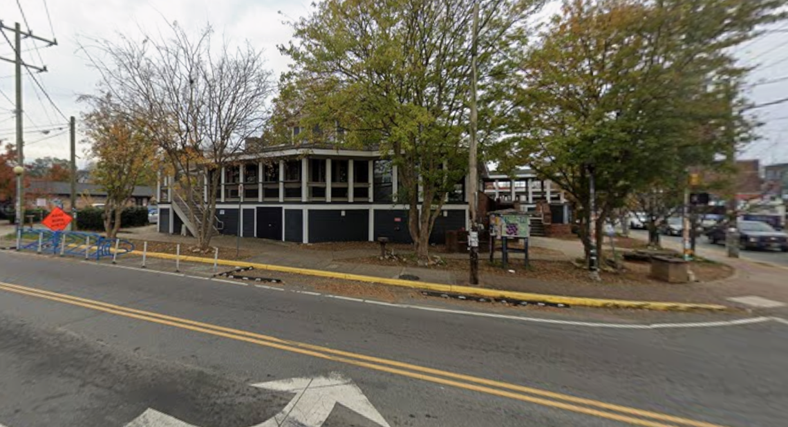 A two-story building with dark siding and an upper-level porch is viewed from across a street in a downtown or village setting. The building is surrounded by trees with fall foliage and sits behind a curb with yellow paint, while cars and other buildings are visible in the background to the right.