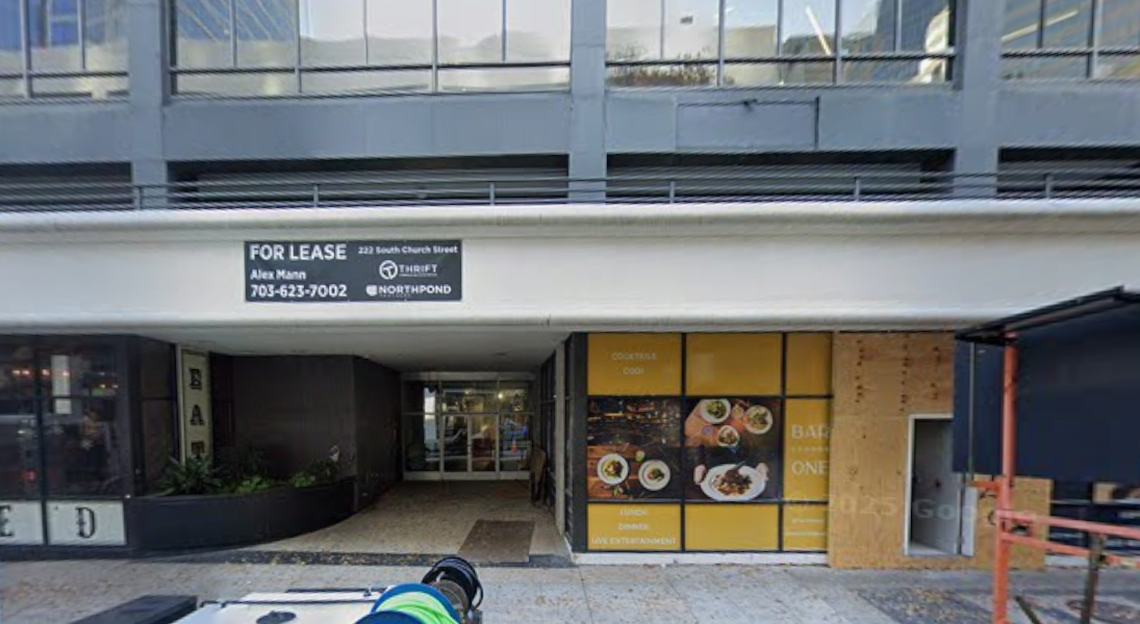 A photo of a street-level commercial space available for lease at 222 South Church Street. A sign above the entrance lists a contact name and phone number. The surrounding storefronts are currently closed or under construction, with yellow advertising posters on the right and a dark interior on the left.