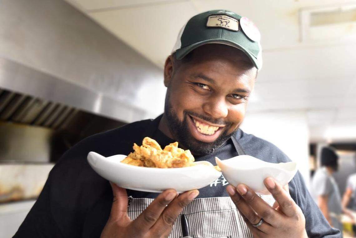 A smiling chef wearing a dark green baseball cap, an apron, and a wedding ring, holds two white, curved bowls of food. The bowl on the left contains pieces of fried chicken, and the bowl on the right holds a white dipping sauce. The chef is looking directly at the camera in a brightly lit commercial kitchen.