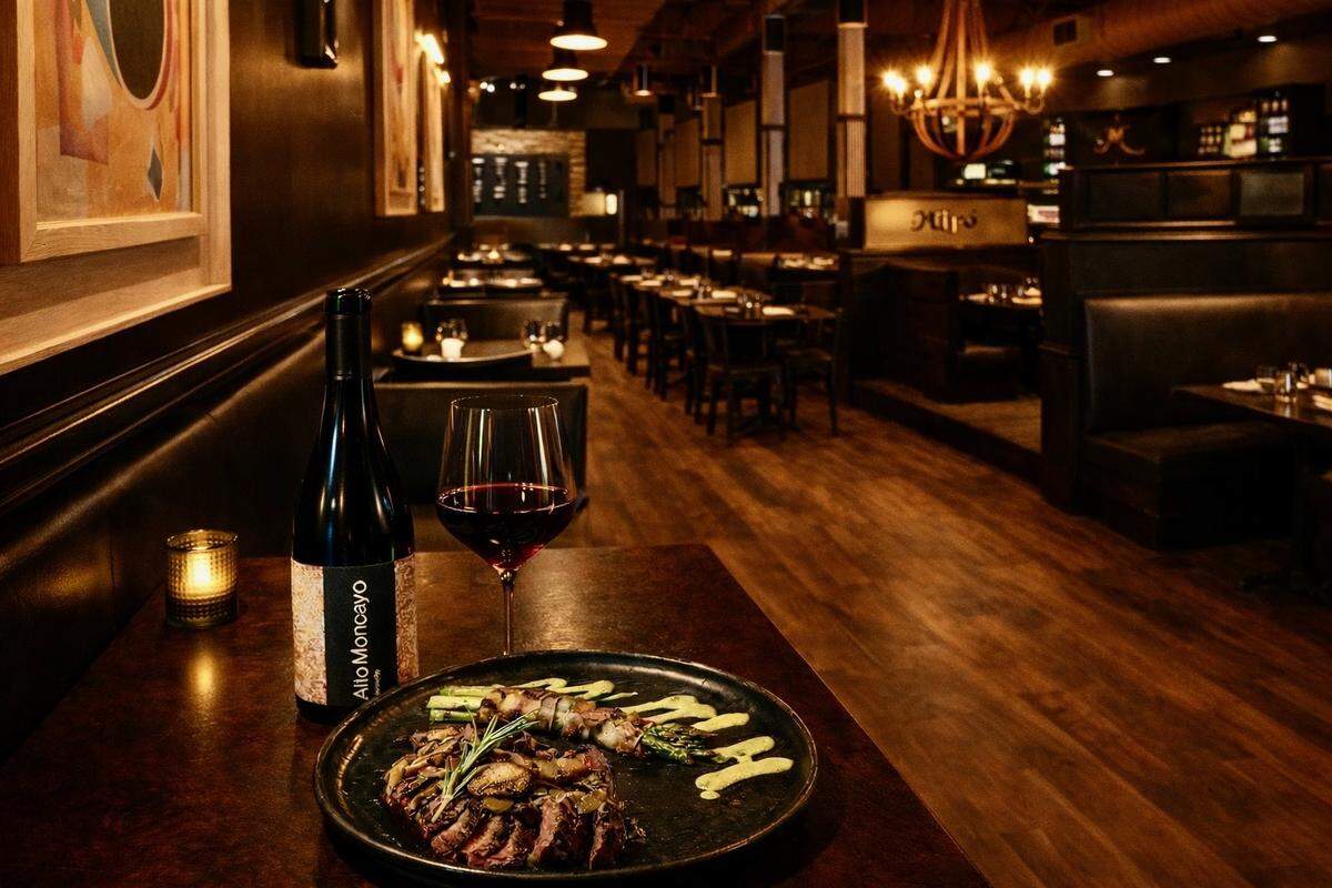In a dimly lit, upscale restaurant with warm wood floors and a glowing chandelier, a plate of sliced sirloin steak topped with mushrooms and rosemary sits on a dark wooden table. Beside the steak is a bottle of Alto Moncayo red wine and a half-filled glass, creating a sophisticated dining atmosphere. The background shows a long, perspective view of the dining room with empty booths, dark walls adorned with framed art, and soft amber lighting.