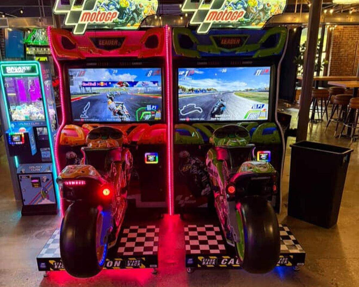Two brightly colored MotoGP motorcycle racing arcade machines stand side-by-side in the dimly lit venue. The games feature checkered platforms and glowing red and green neon accents, with the screens showing a first-person view of a racetrack.