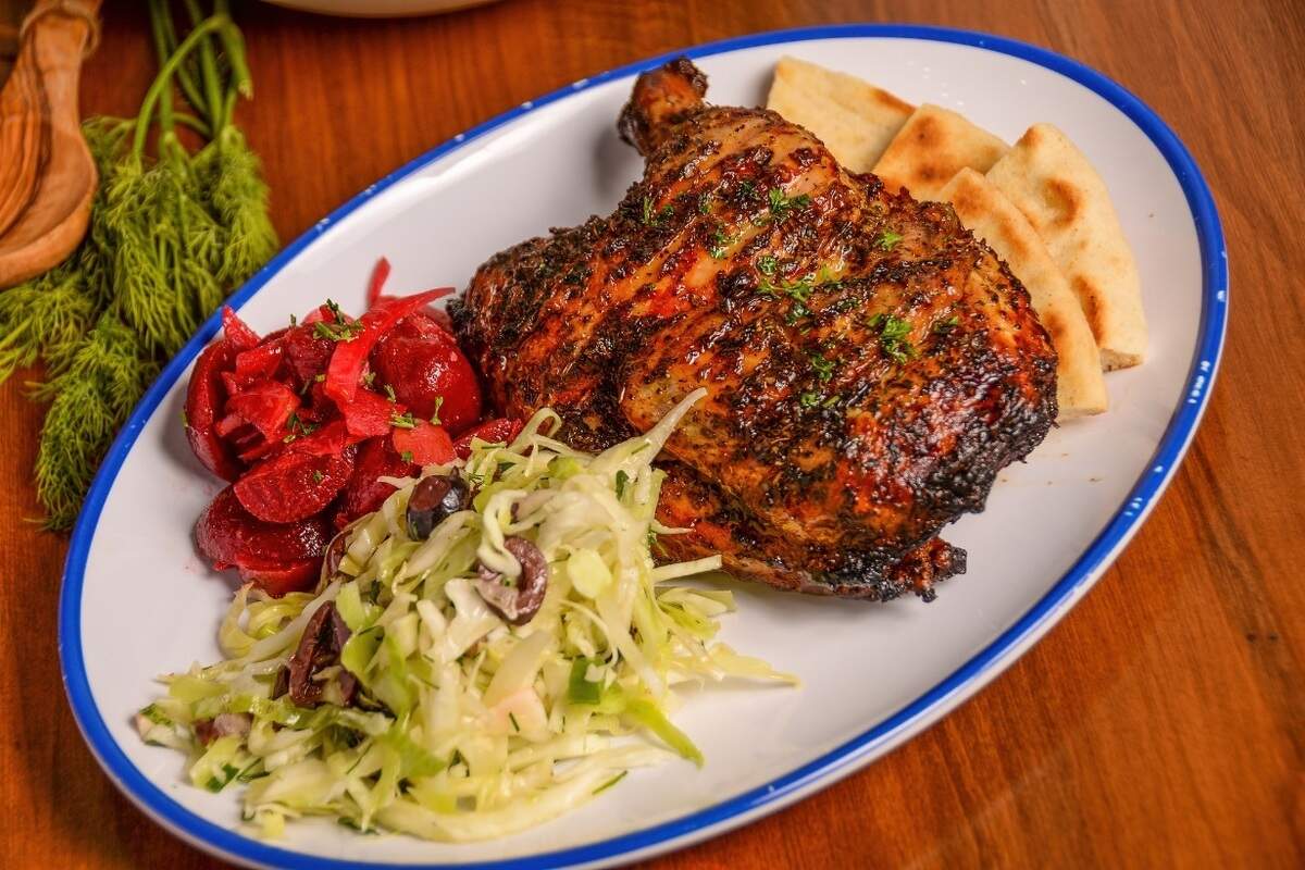 A roasted, seasoned half chicken with a caramelized, herb-dusted skin takes center stage on an oval plate with a blue border. It is accompanied by a mound of shredded cabbage slaw mixed with sliced olives, a side of chopped red beets, and triangles of pita bread. The plate rests on a warm wooden surface next to fresh green dill sprigs and wooden serving utensils.