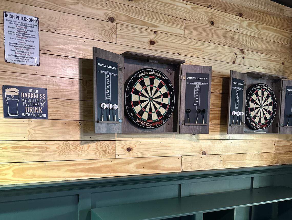 Multiple professional dartboard cabinets are mounted on a light-colored wood-plank wall. Each cabinet includes a scoreboard and slots for holding darts, situated near humorous wall signs about drinking and “Irish Philosophy.”