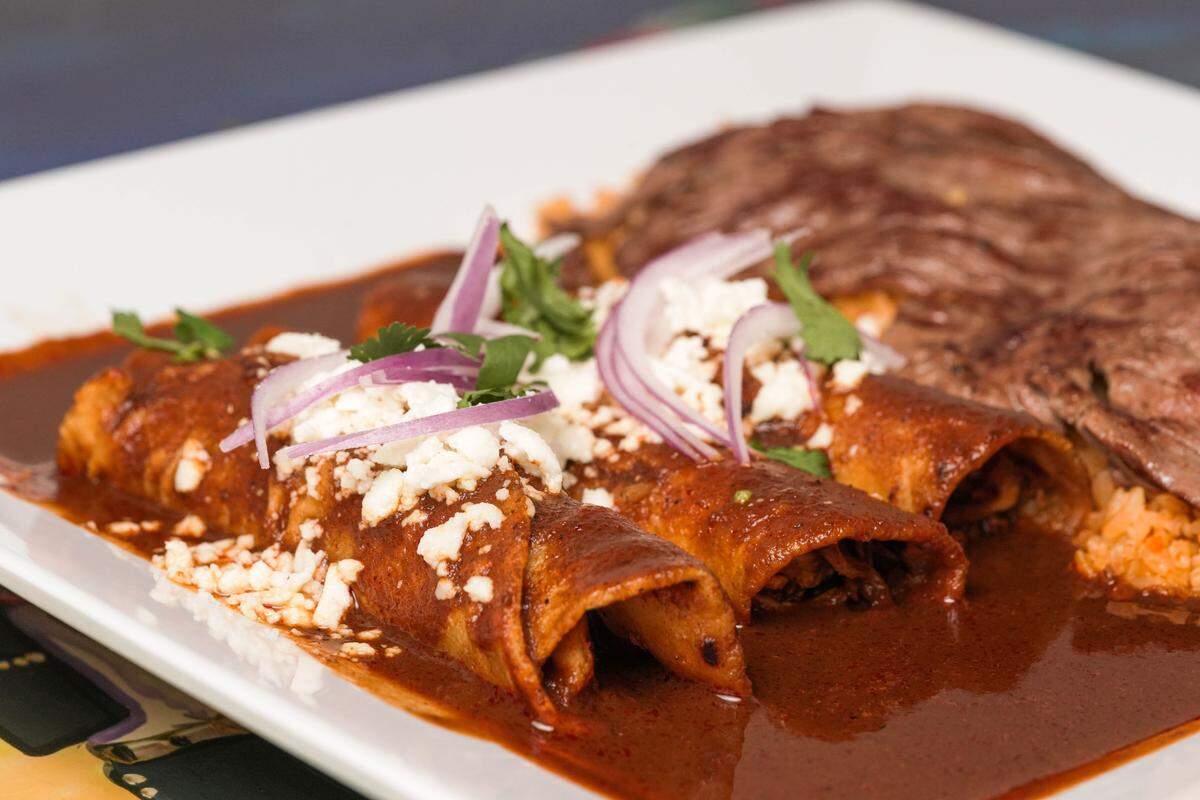 Three rolled enchiladas are drenched in a rich, dark brown mole sauce and garnished with crumbled white cheese and slivers of red onion. In the background, a large piece of grilled skirt steak sits next to a small mound of Mexican rice. The meal is served on a clean white rectangular plate, highlighting the deep colors of the sauce.
