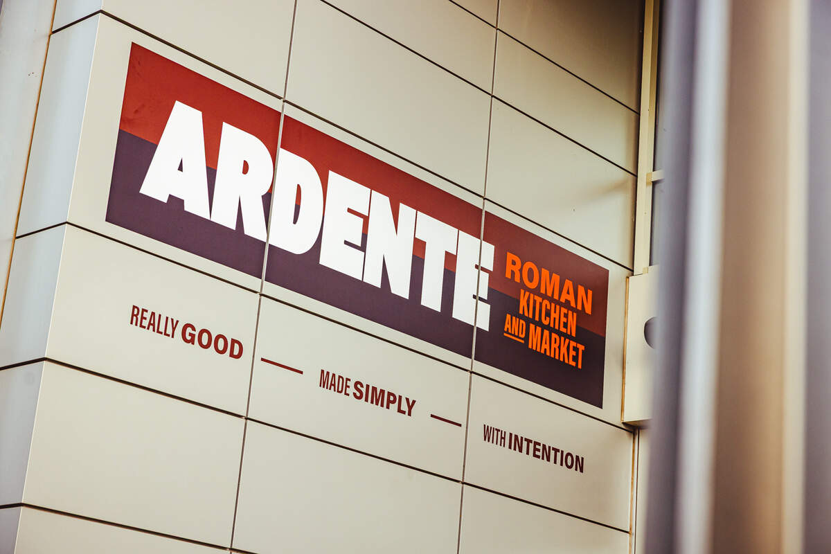 An outdoor sign on a tiled, light-cream building facade reads “ARDENTE ROMAN KITCHEN AND MARKET” in bold white and orange lettering against a dark rectangular background. Below the main name, the restaurant’s tagline is written in smaller, spaced-out capital letters: “REALLY GOOD — MADE SIMPLY — WITH INTENTION.” The shot is taken from a low angle, looking slightly upward at the exterior branding.