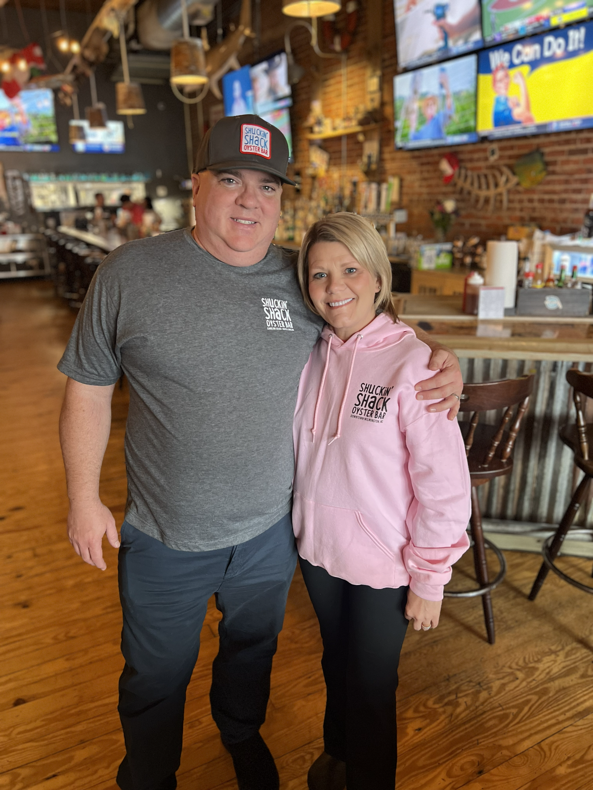 A man and a woman stand close together smiling inside a Shuckin’ Shack restaurant. The man wears a grey branded t-shirt and a matching hat, while the woman wears a light pink branded hoodie, with a brick wall and sports bar seating behind them.