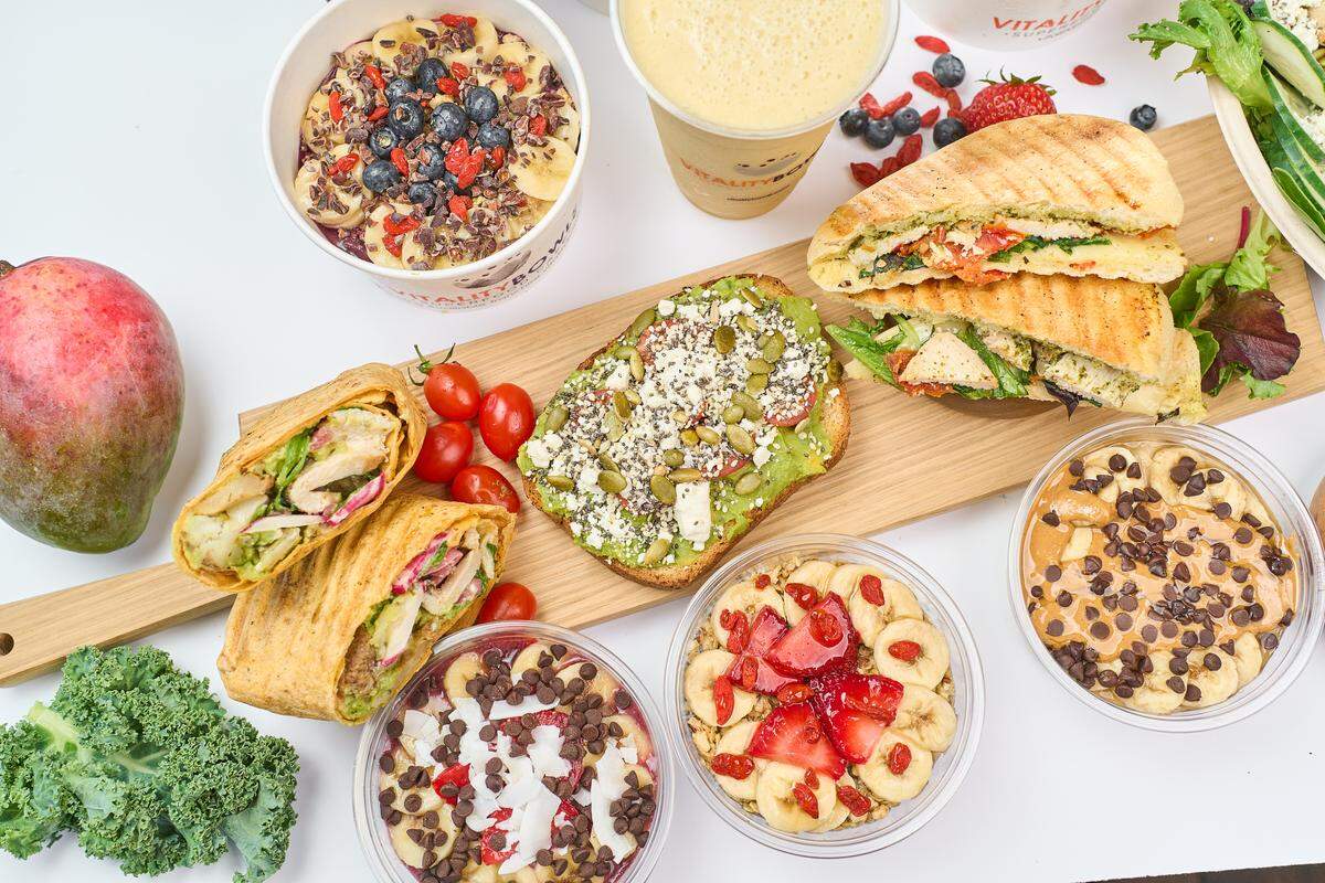 A top-down view of a wooden serving board featuring a chicken wrap, avocado toast with feta and seeds, and a pesto panini. Surrounding the board are several colorful açaí bowls topped with fruit, chocolate chips, and coconut shavings.