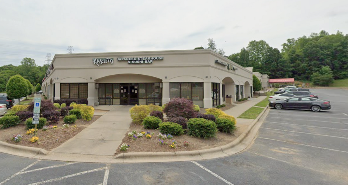 This is a daytime, outdoor shot of the exterior storefront for Kabuto Japanese Steakhouse & Sushi Bar, located in a light beige strip mall building. A concrete walkway leads up to the restaurant’s glass entrance doors, flanked by landscaped garden beds filled with green and purple shrubs and small colorful flowers. To the right, an asphalt parking lot contains several parked cars, with a dense line of green trees filling the background under an overcast sky.