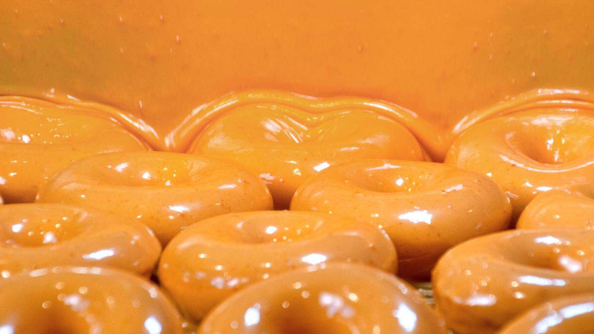A wide view captures a row of donuts passing under a continuous, “waterfall” curtain of thick, bright orange glaze. The liquid coating perfectly envelopes each donut as they move along the conveyor belt in a commercial bakery setting. The scene is saturated with warm orange tones, emphasizing the smooth and gooey texture of the icing.