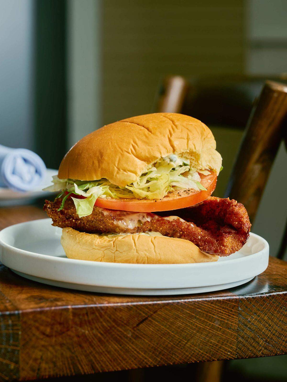 A crispy, cornmeal-breaded fish fillet is served on a soft brioche bun with a thick tomato slice, shredded lettuce, and creamy tartar sauce. The sandwich is presented on a white plate atop a rustic wooden table in a brightly lit dining setting.