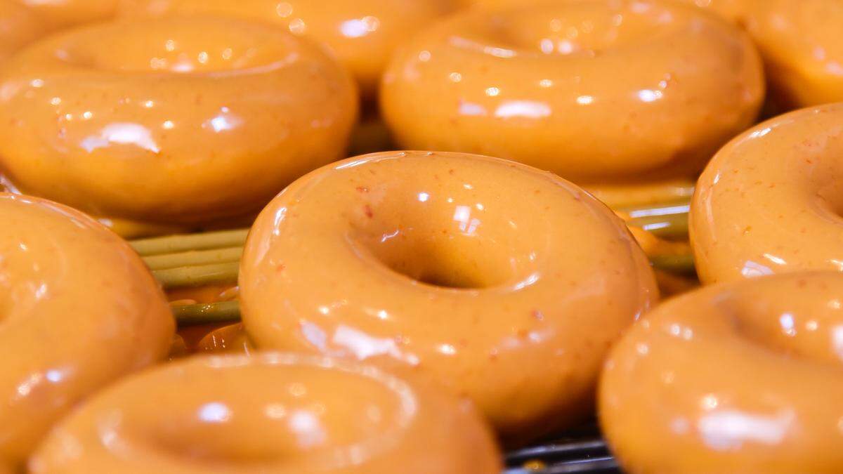 A close-up, shallow-depth-of-field shot highlights several donuts covered in a vibrant, glossy orange glaze. The foreground donut is in sharp focus, revealing tiny orange flecks within the smooth, reflective coating. They are arranged in neat rows on a metal cooling rack, suggesting they are fresh from the glazing line.