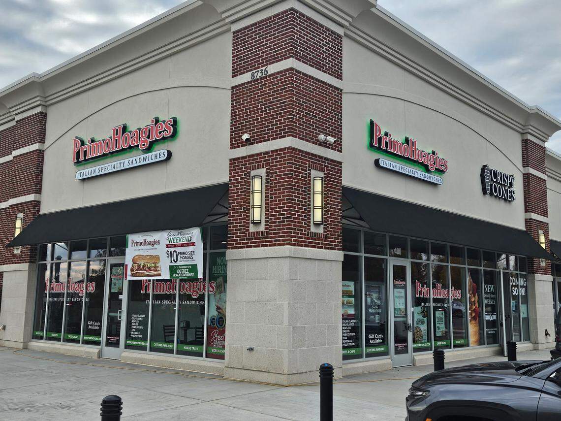 An exterior photo of a PrimoHoagies storefront located in a modern strip mall. The building has beige stucco and red brick accents. The green and white “PrimoHoagies Italian Specialty Sandwiches” sign is visible above the dark awning. A promotional sign is taped to the window, and an adjacent storefront, “Crispy Cones,” is visible to the right.