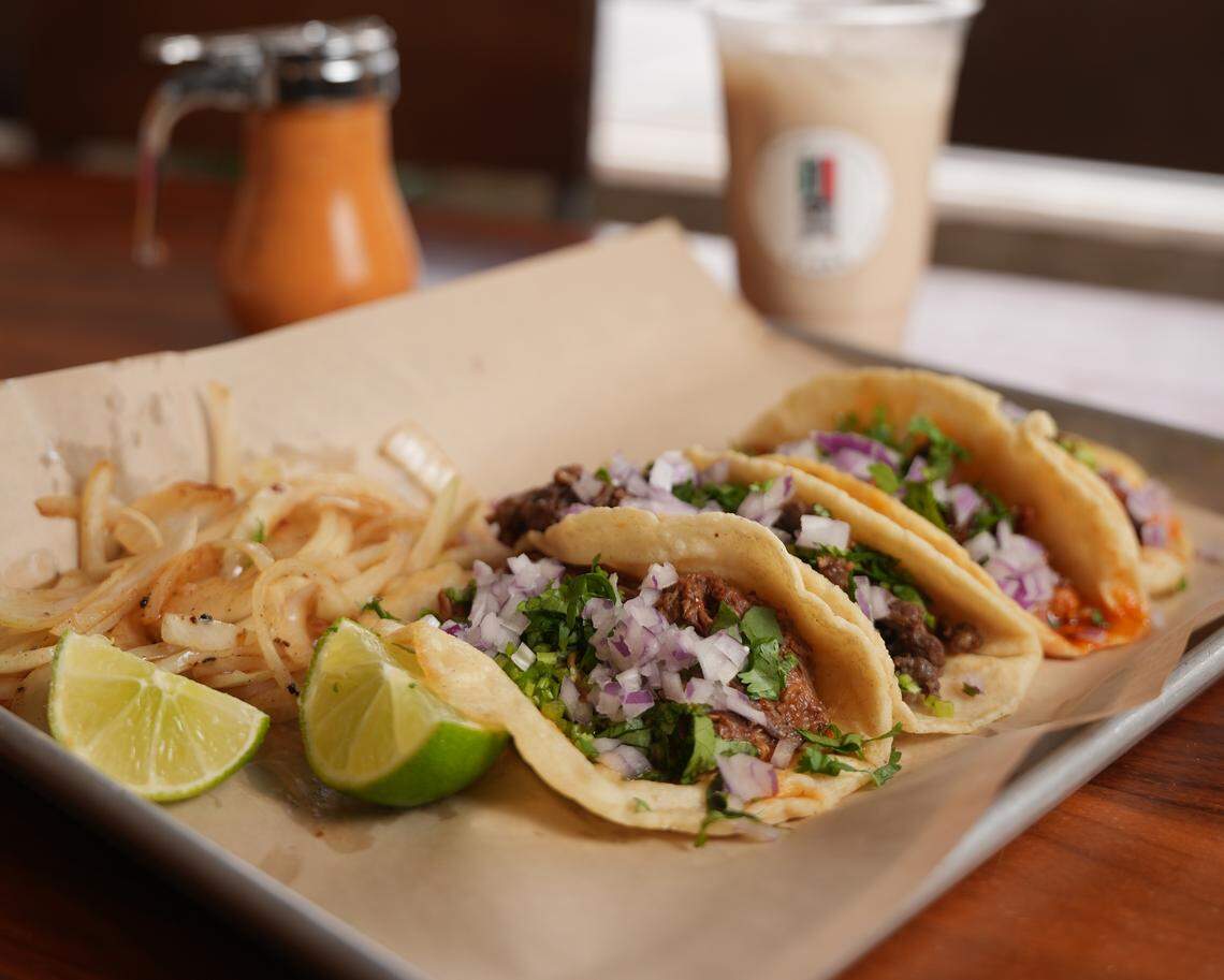 Three tacos with meat, cilantro, and diced red onion are arranged on a piece of paper on a metal tray, accompanied by a pile of sautéed white onions and two lime wedges. In the background, there is a small jar of orange sauce and a blurred plastic cup containing a light-colored beverage.
