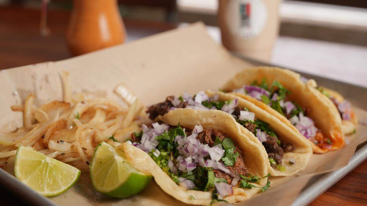 Three tacos with meat, cilantro, and diced red onion are arranged on a piece of paper on a metal tray, accompanied by a pile of sautéed white onions and two lime wedges. In the background, there is a small jar of orange sauce and a blurred plastic cup containing a light-colored beverage.