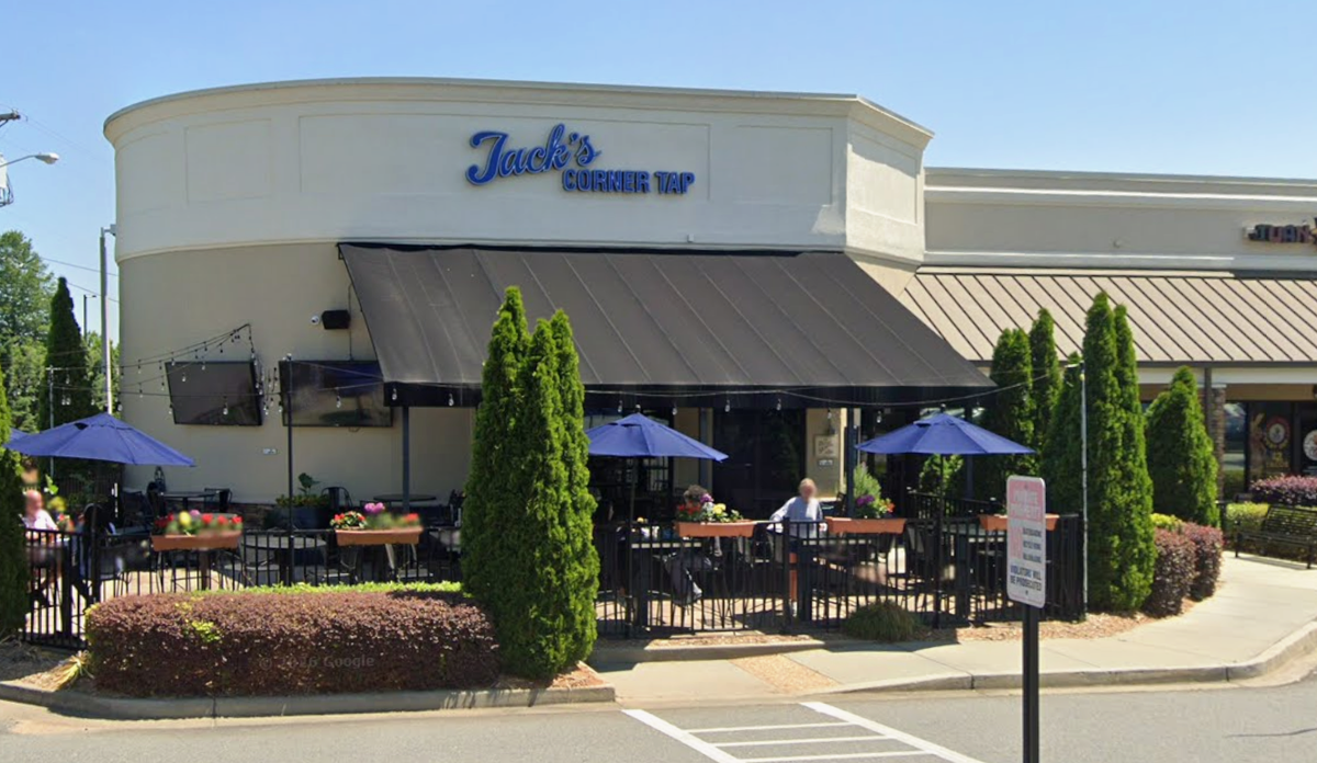 This image shows the exterior of Jack’s Corner Tap, a neighborhood restaurant featuring a rounded cream-colored facade and a large black awning. A gated outdoor patio is filled with black tables, blue umbrellas, and wooden planter boxes overflowing with colorful flowers. Tall, neatly trimmed evergreen trees and manicured hedges line the perimeter of the seating area under a clear blue sky.