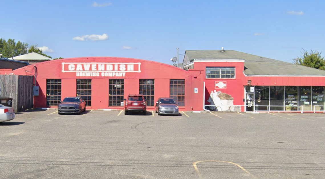 The exterior of Cavendish Brewing Company is a long, bright red building with a curved roofline and several large, multi-paned windows. A paved parking lot sits in front of the building under a clear blue sky, with a few cars parked near the entrance.