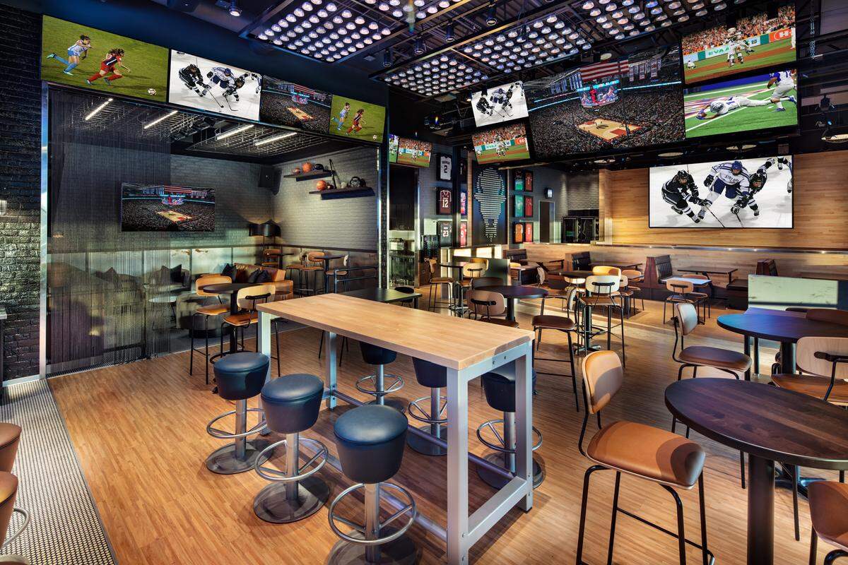 The interior of a Buffalo Wild Wings restaurant features a modern sports bar layout with numerous high-definition televisions mounted along the walls and ceiling. Various seating options are visible, including wooden communal tables with blue barstools and smaller circular tables paired with tan chairs.