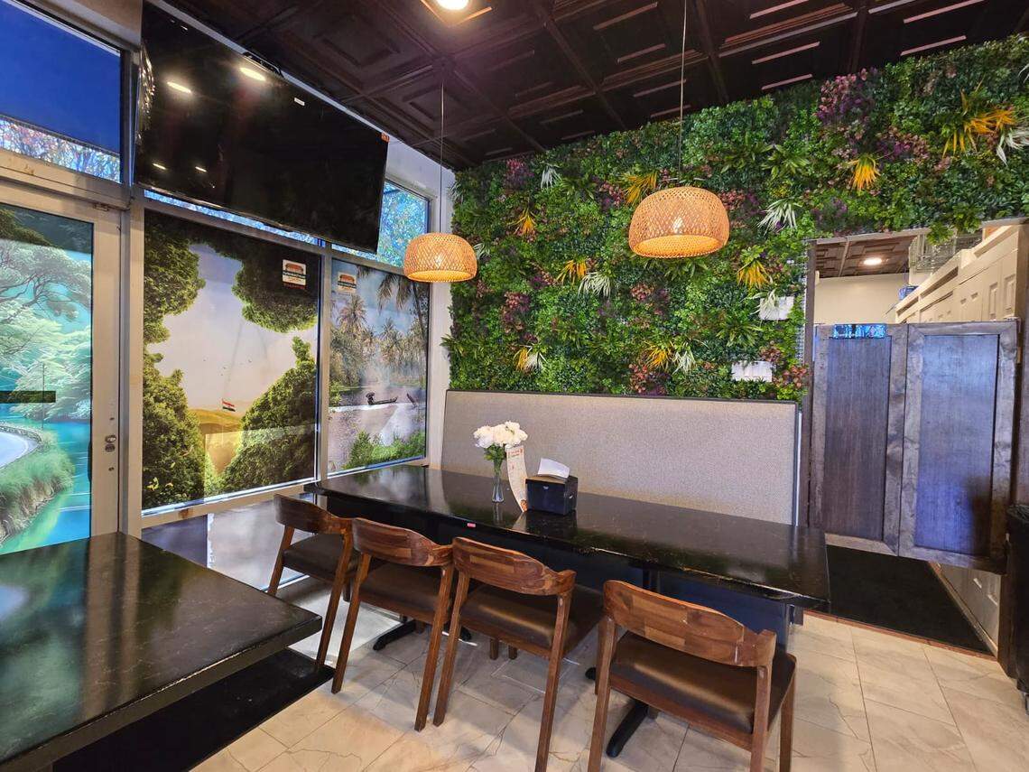 A booth-style dining area with a long black table and four wooden chairs sits against a textured wall covered in artificial greenery. The area is lit by two woven basket pendant lamps and large window graphics depicting lush landscapes.