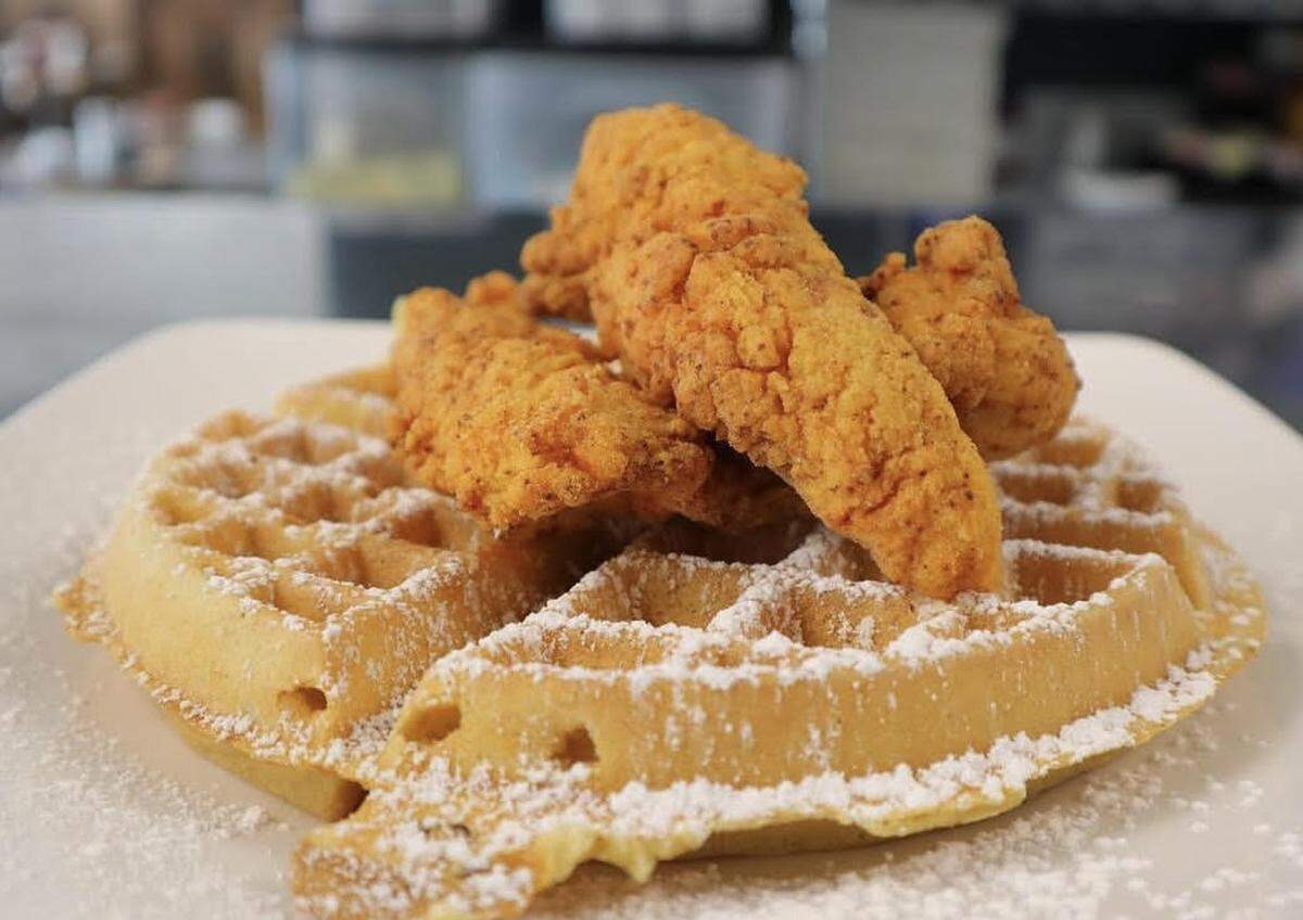 A plate of chicken and waffles is presented in a close-up, appetizing view. Three crispy, golden-fried chicken tenders are stacked neatly on top of a fluffy, Belgian-style waffle. The entire dish is lightly dusted with a fine layer of powdered sugar.