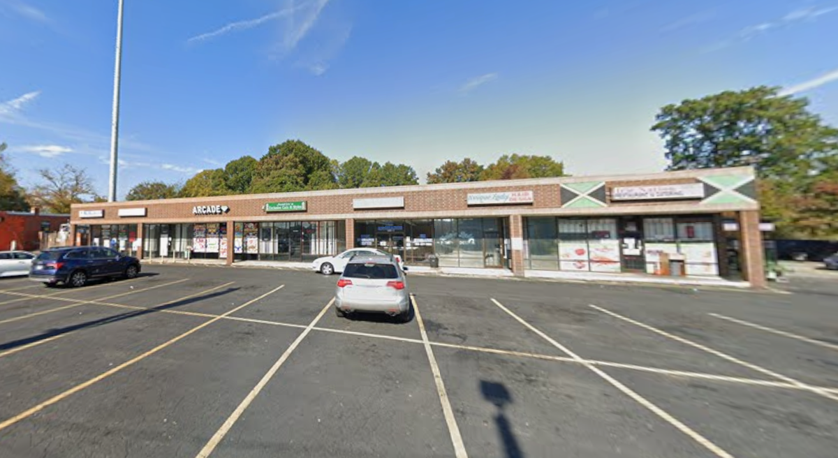A wide, exterior view of a single-story, brick strip mall with a large, mostly empty asphalt parking lot in the foreground. Several small businesses are visible, including “ARCADE,” a Jamaican flag design on one storefront, and “Dye Natural Hair.” The sky is blue and partially cloudy, and there are trees with green and autumn foliage in the background. Several cars are parked in the lot.