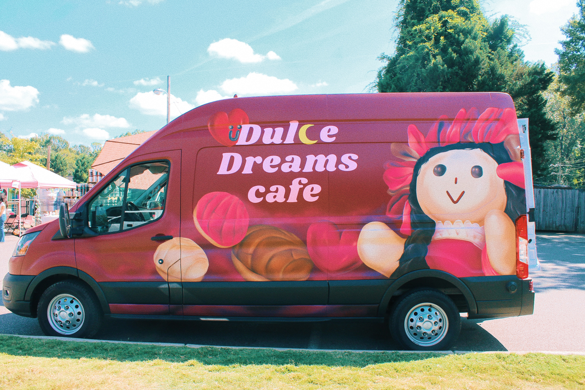 A bright maroon food service van for “Dulce Dreams cafe,” featuring a large graphic of a smiling, traditional Mexican doll and several pieces of concha-style sweet bread on the side panel. The van is parked on grass next to a paved area under a sunny, light blue sky.