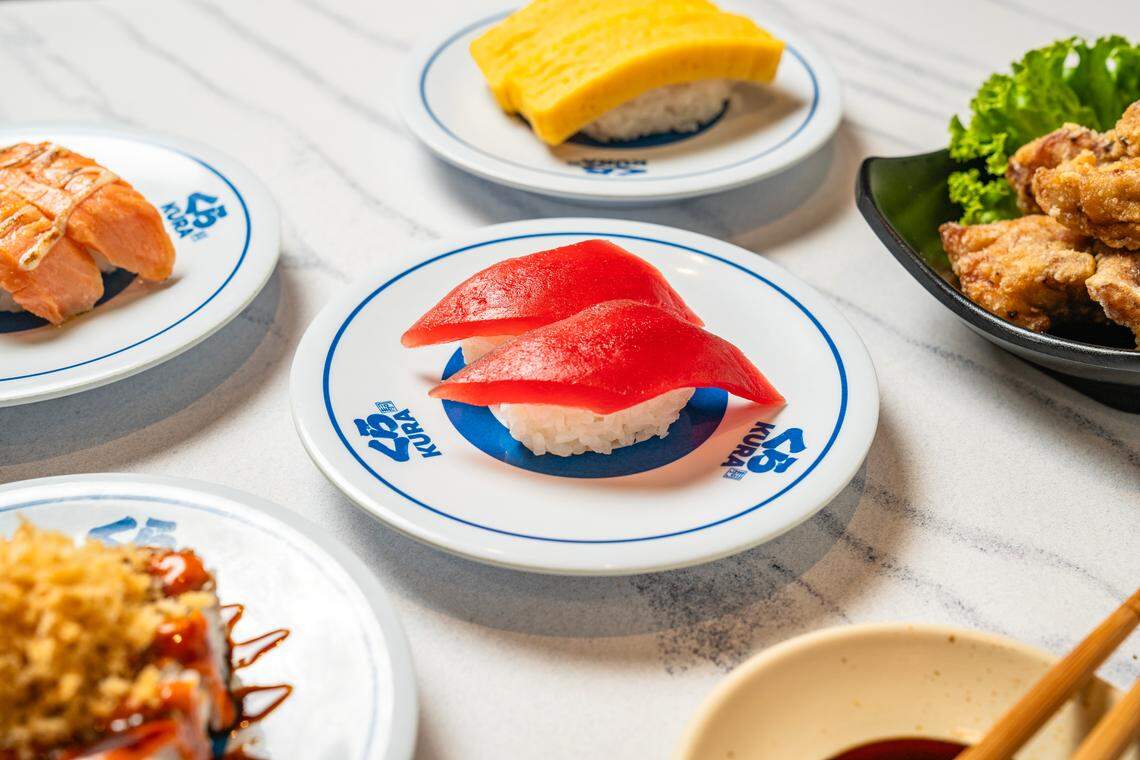 A close-up shot focuses on two pieces of bright red tuna nigiri served on a white and blue branded plate. In the background, other dishes including tamago sushi, seared salmon, and a bowl of fried karaage chicken are scattered across a light marble tabletop. The composition uses a shallow depth of field to highlight the fresh texture of the tuna.
