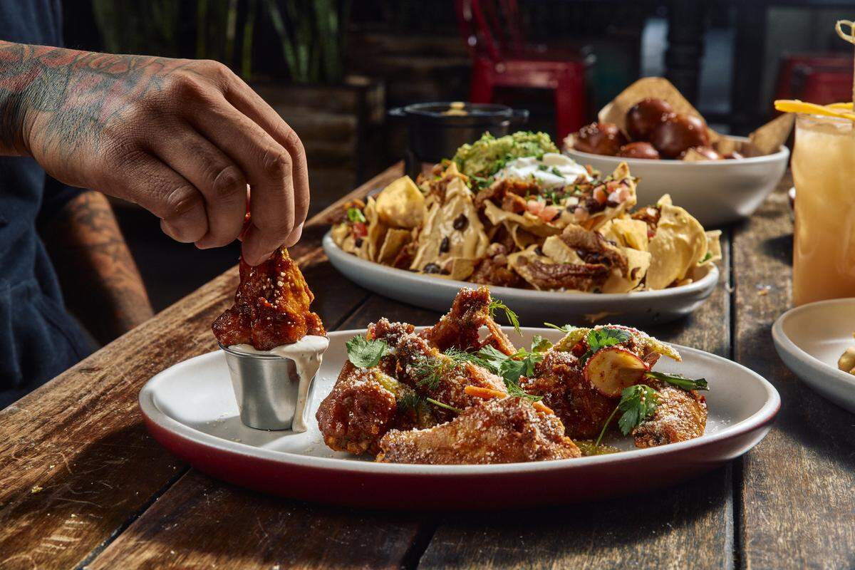 A close-up shot of a person dipping a crispy, glazed chicken wing into a small metal cup of creamy ranch dressing. In the background, plates of nachos and pretzel bites sit on the rustic wooden table, blurred to keep the focus on the wings.