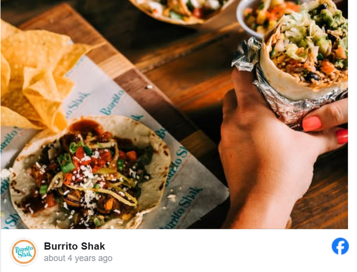 A close-up shot features a hand holding a large, foil-wrapped burrito filled with rice, black beans, lettuce, and guacamole. To the left, an open-faced taco topped with seasoned meat, fresh cilantro, and crumbled cheese sits on branded wax paper alongside a pile of tortilla chips. The wooden table and warm lighting create a rustic, inviting atmosphere for a “Burrito Shak” meal.