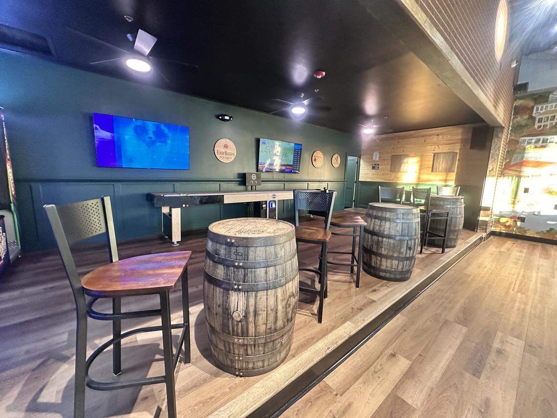 The interior of the bar features a rustic-industrial theme with teal walls, wood floors, and whiskey barrels used as cocktail tables. A long shuffleboard table sits against one wall, while flat-screen TVs and a prize crane machine are visible in the background.