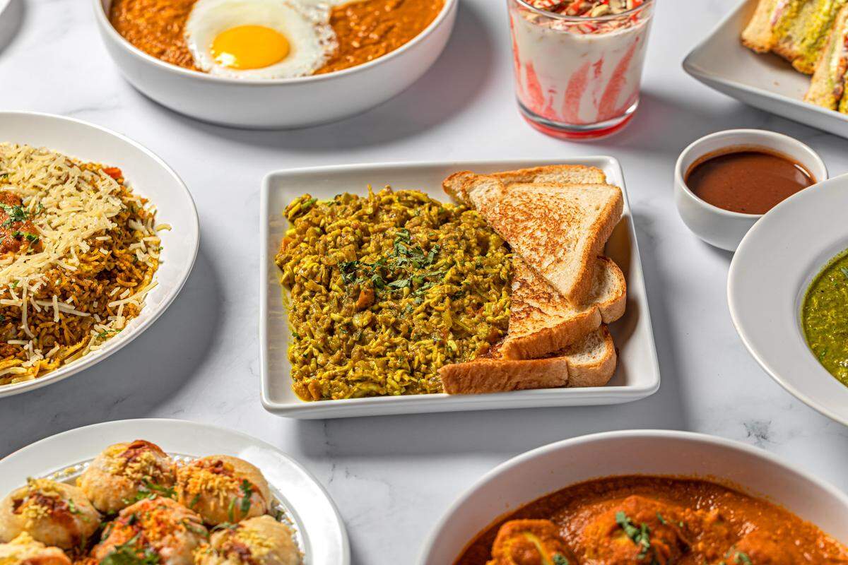 A close-up of a spread of Indian breakfast and lunch dishes on a white marble surface, centered on a square plate of vibrant green spiced scrambled eggs, or Anda Bhurji, served with white toast. Surrounding dishes include biryani, a yogurt drink, a bowl of curry topped with a fried egg, and savory fried snacks.