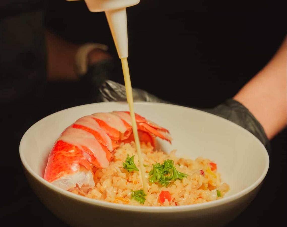 A chef wearing black gloves pours a steady stream of creamy yellow sauce from a squeeze bottle onto a lobster tail. The lobster sits atop a bed of seasoned rice mixed with herbs and vegetables in a clean white bowl. The dark, blurred background focuses all attention on the vibrant colors of the seafood and the drizzling sauce.