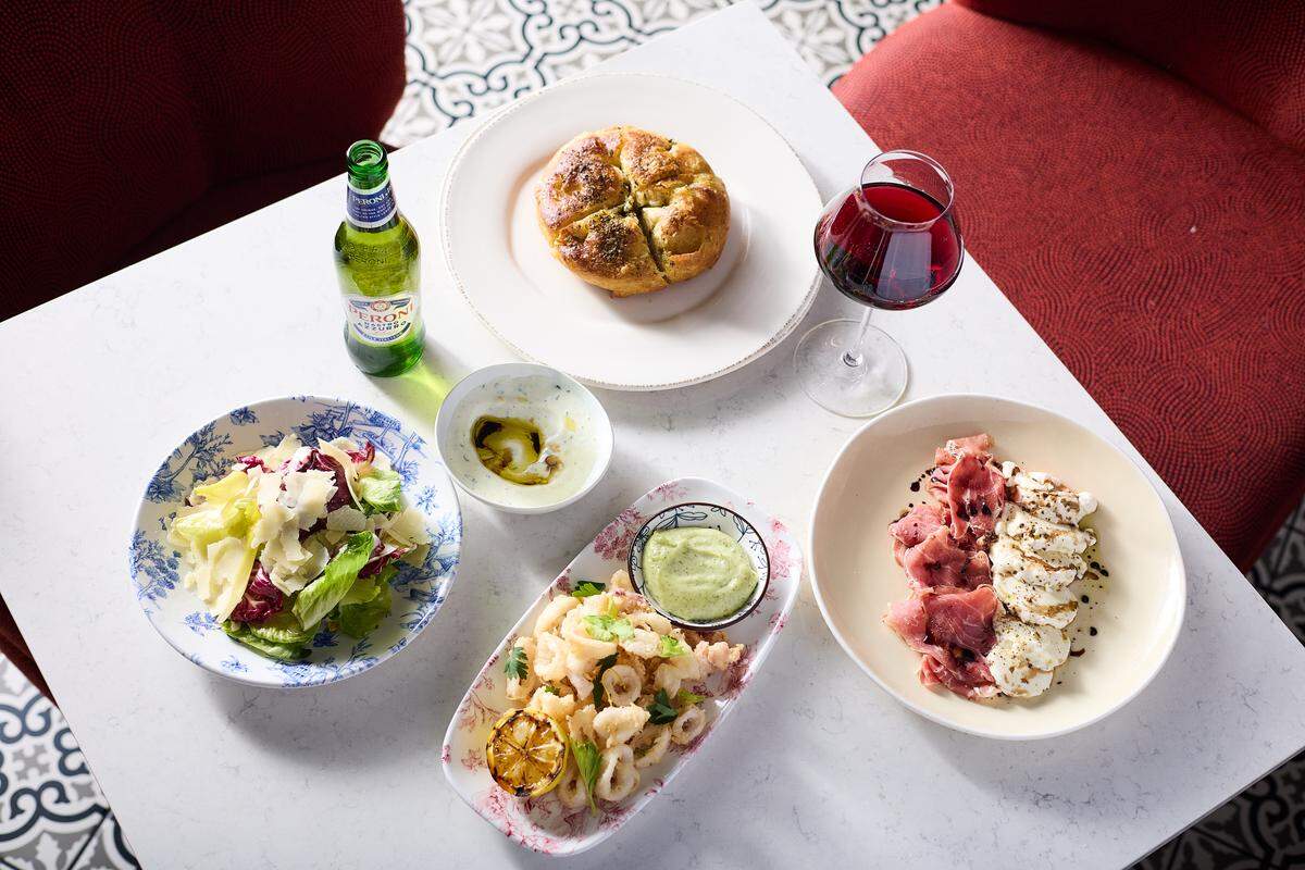 A high-angle view shows an assortment of Italian appetizers spread across a white marble table. The spread includes a whole round focaccia, a plate of prosciutto and mozzarella, fried calamari with a grilled lemon, and a fresh green salad with shaved parmesan. A bottle of Peroni beer and a glass of red wine sit among the dishes, creating an inviting lunch scene.