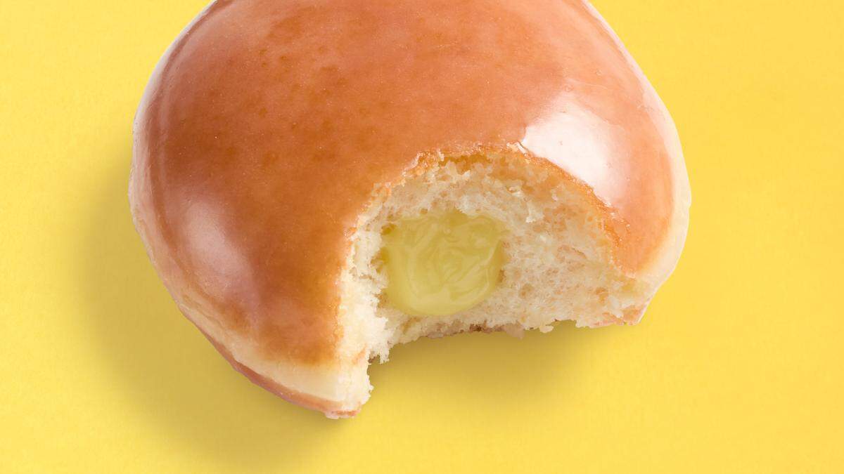 A close-up shot features a round, glazed doughnut with a bite taken out of it, centered against a solid yellow background. The glossy, golden-brown glaze catches the light, revealing a soft, fluffy interior filled with a bright yellow lemon curd. The missing bite showcases the gooey texture of the filling nestled perfectly inside the pastry.