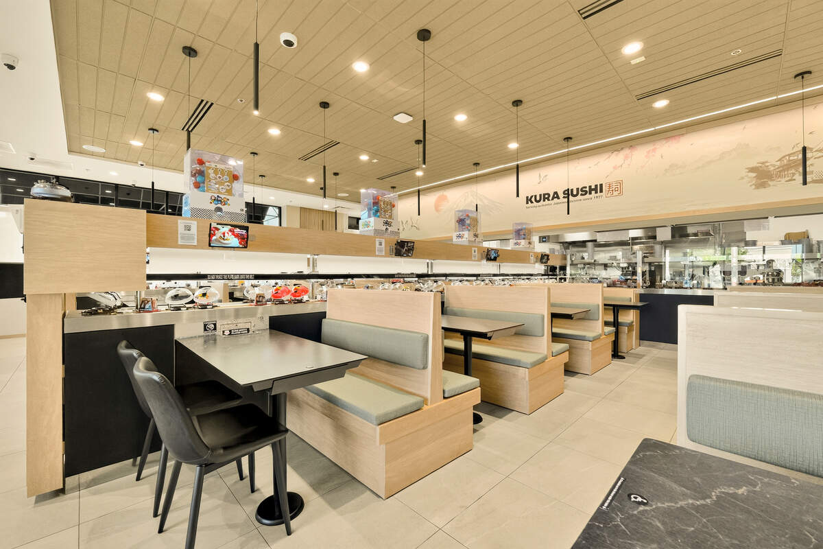 The bright interior of a Kura Sushi restaurant features rows of light wood booths with pale green cushions alongside a multi-tiered conveyor belt system. Small, white capsule toy dispensers sit atop the conveyor tracks, which stretch across the dining room toward an open kitchen area in the back. A wall mural with Japanese-inspired artwork and the “Kura Sushi” logo decorates the space beneath a grid of modern pendant lighting.