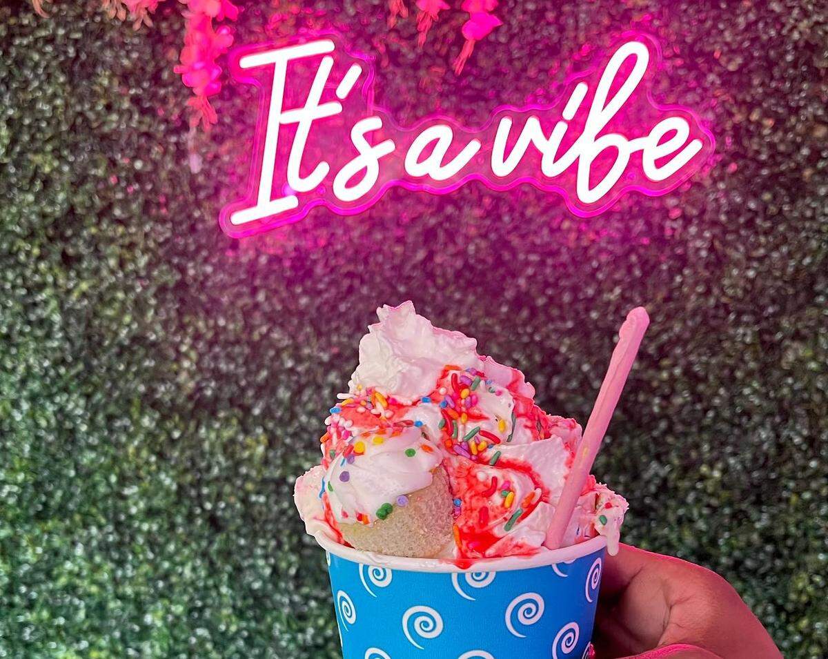 A festive ice cream sundae in a blue cup is topped with whipped cream, red syrup, and colorful sprinkles, and is held in front of an artificial green hedge wall. Above the sundae, a bright pink neon sign cursive reads, “It’s a vibe.”