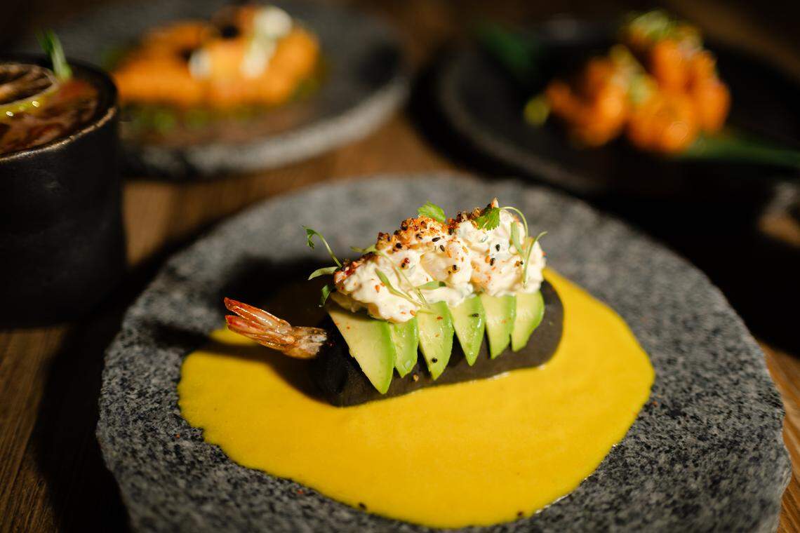 A close-up shot of a sophisticated Peruvian dish, the Causa Panko starter, is centered on a textured gray plate. The dish features a bed of creamy yellow sauce topped with a dark mound, slices of avocado, and a generous dollop of white cream and seasonings, with a shrimp tail visible at the side.