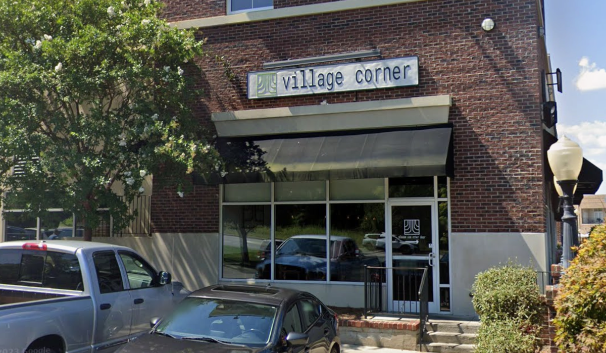 The exterior of Village Corner features a brick facade with a black awning and a large sign above the entrance. Several cars are parked on the street in front of the building, which is framed by a white-flowering tree to the left.