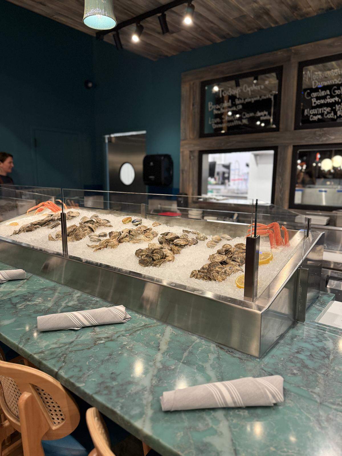 A sleek oyster bar features various shellfish and crab legs displayed on a bed of crushed ice behind a glass partition. The setting is modern with a teal wall, wooden accents, and a marble-patterned countertop set with napkins.