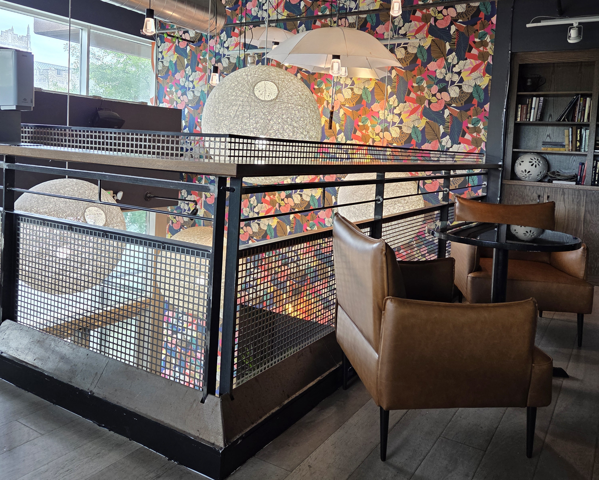 This photo looks across an upstairs mezzanine with a black metal railing overlooking the main floor below. The ceiling is industrial with exposed silver ductwork, and the space is decorated with large, unique lighting fixtures, including two shaped like white umbrellas.